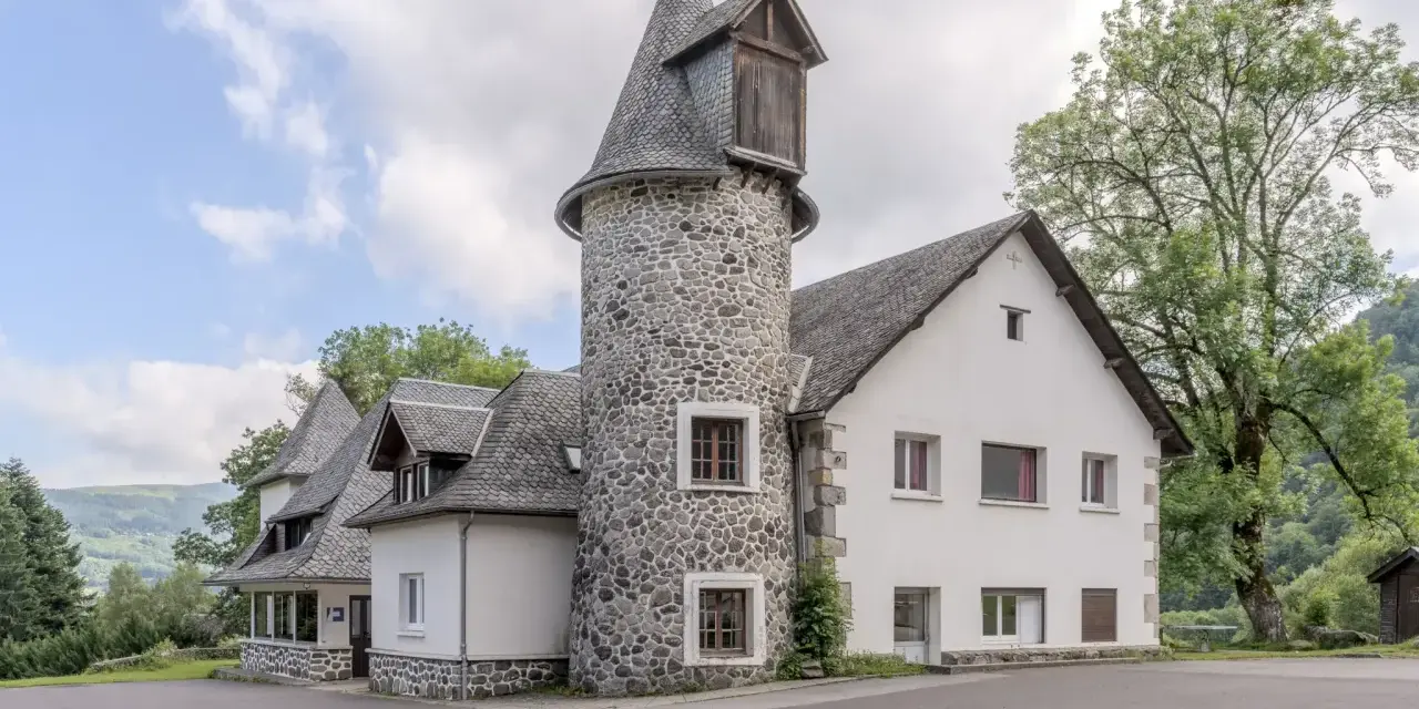 Bâtiment blanc avec une tour ronde en pierre et un toit en ardoise, entouré d'arbres et de verdure.