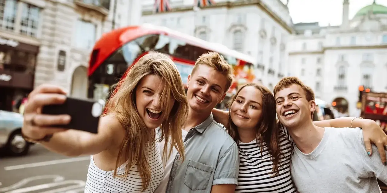 Quatre jeunes souriants prennent un selfie à Londres, avec un bus rouge et des drapeaux britanniques en arrière-plan.