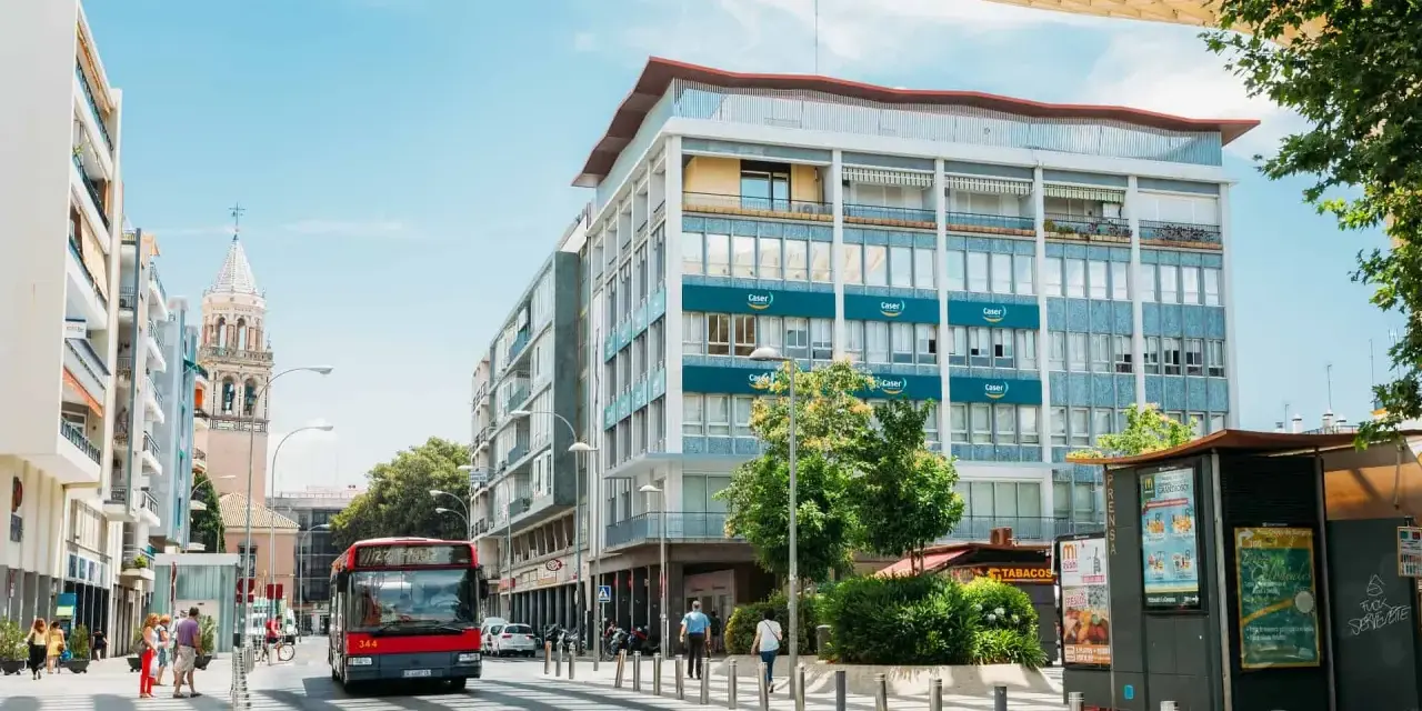 Un bus rouge circule dans une rue bordée d'immeubles et d'un clocher, sous un ciel bleu.