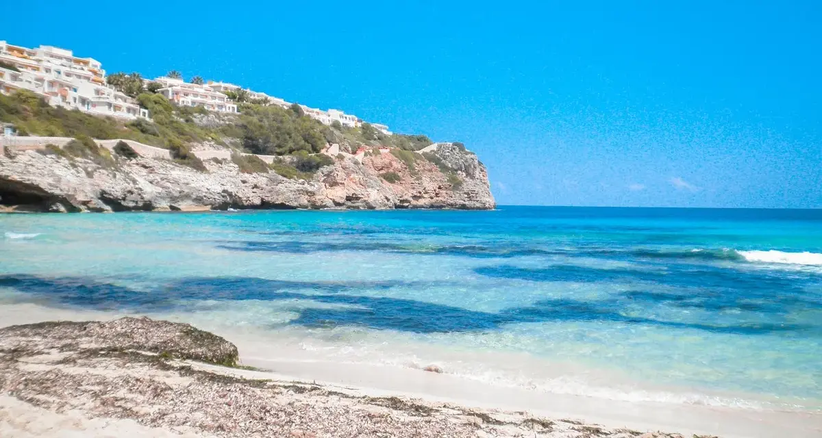 Plage de Majorque avec eaux turquoises, falaises et bâtiments blancs à flanc de colline.
