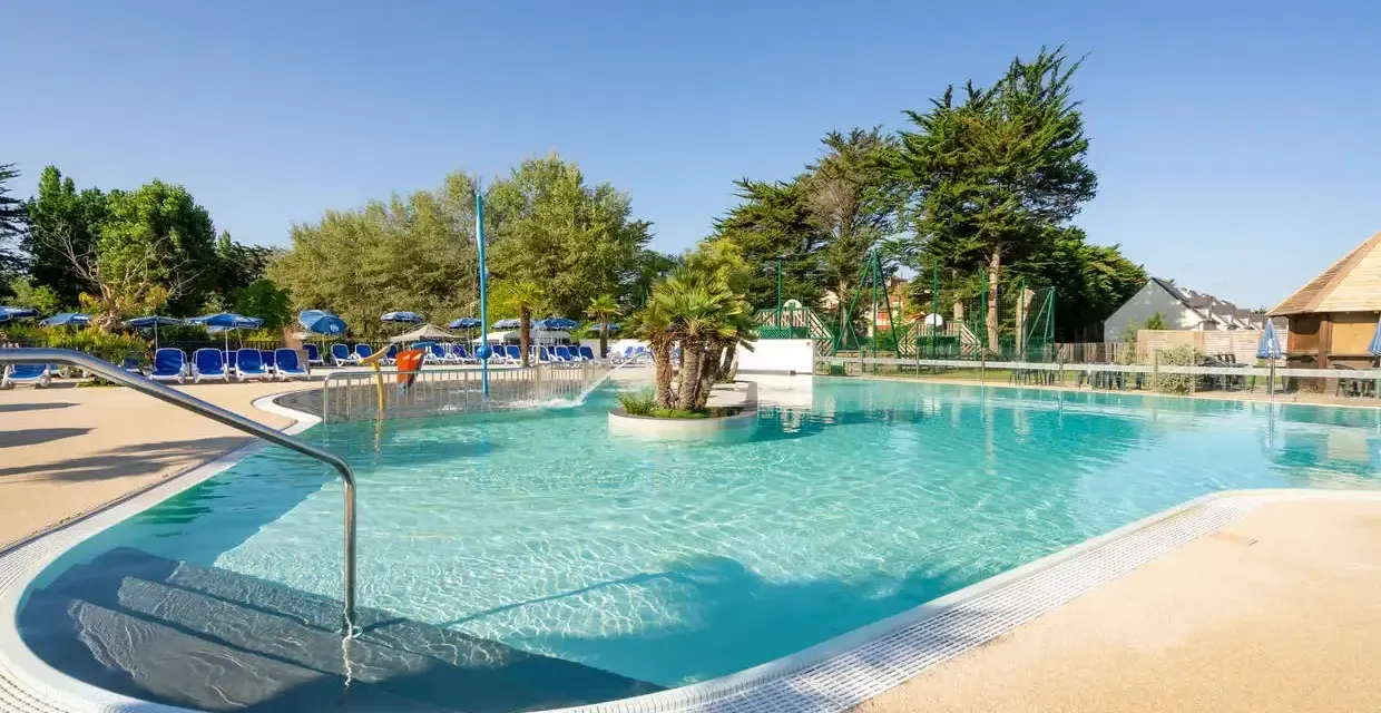 Piscine extérieure avec chaises longues bleues, parasols et végétation luxuriante sous un ciel bleu clair.