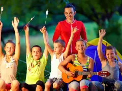 Une animatrice de colonie de vacances à l'étranger supervise des enfants joyeux autour d'un feu de camp, certains grillant des marshmallows, d'autres jouant de la guitare.