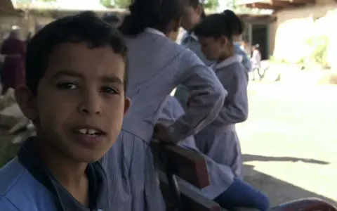 Un garçon souriant pose devant des filles en uniforme dans la même cour d'école.