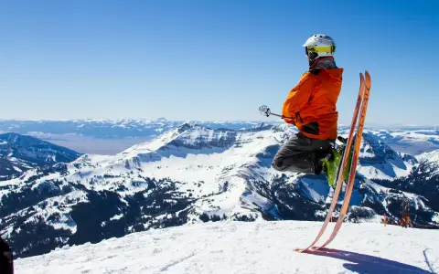 Un skieur en l'air, prêt à dévaler la pente. Il porte un casque et une veste orange vif, rappelant les règles pour skier en toute sécurité. Le paysage montagneux enneigé est spectaculaire.
