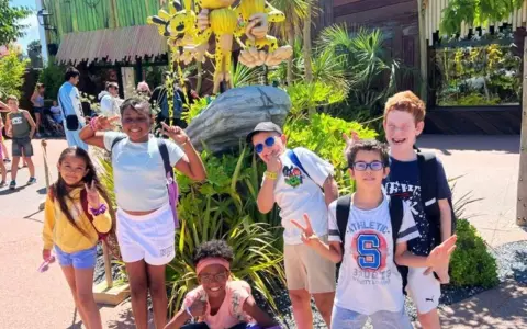 Des enfants joyeux font des signes de paix devant une attraction, un avis "supernova juniors" est visible.