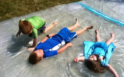 Trois enfants s'amusent sur un jeu d'eau pour kermesse, glissant sur une bâche mouillée sous le soleil.