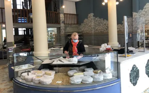 Une fromagère masquée prépare des fromages dans une vitrine réfrigérée.