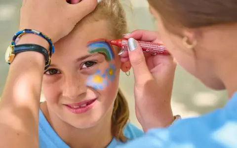 Une petite fille souriante se fait maquiller sur la joue un arc-en-ciel et des fleurs, au stand maquillage d'une kermesse.