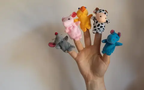 Main d'une personne avec cinq marionnettes à doigts en feutrine : souris grise, lapin rose, canard jaune, vache noire et blanche, hippocampe bleu.