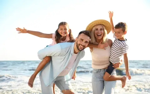 Famille souriante sur la plage, père portant sa fille sur le dos, mère tenant son fils.