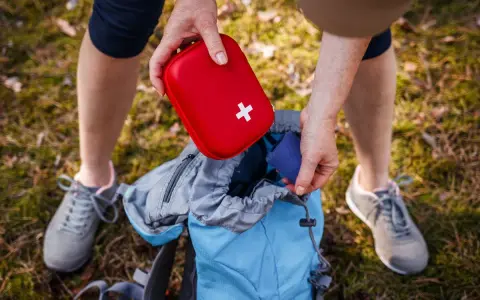 Une personne prépare son sac à dos pour une randonnée, rangeant une trousse à pharmacie enfant rouge avec une croix blanche.