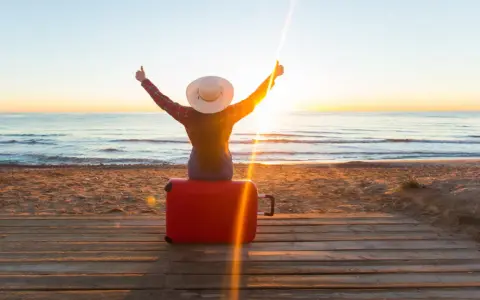 Personne assise sur une valise rouge sur une plage au coucher du soleil, bras levés en signe de victoire.