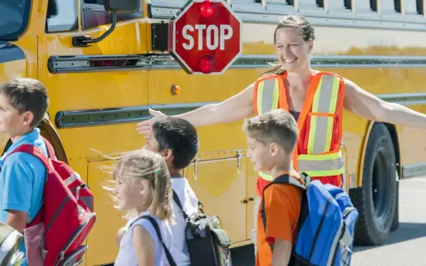 Des enfants montent dans un bus scolaire jaune, guidés par une adulte portant un gilet de sécurité orange.