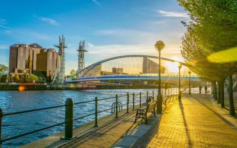 Promenade ensoleillée au bord de l'eau, avec un pont moderne et des bâtiments reflétant le soleil. Idéal pour visiter Manchester ou Liverpool.