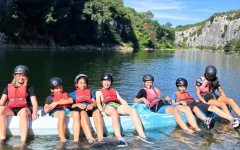 Des enfants en gilets de sauvetage et casques sur un raft bleu sur une rivière, entourés de verdure et de falaises.