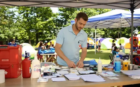 Un homme en polo bleu prépare du matériel médical sur une table, avec des tentes et des enfants en arrière-plan.