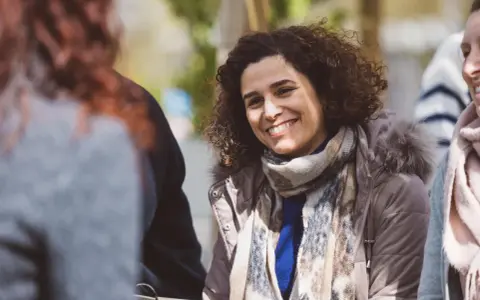 Une femme souriante aux cheveux bouclés porte une écharpe et une veste à capuche en fourrure.