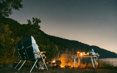 Personne assise près d'un feu de camp sous un ciel étoilé, avec une table de camping et un lac.