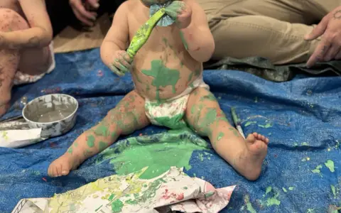 Bébé couvert de peinture verte, tenant un pinceau, assis sur une bâche bleue.