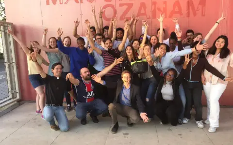 Un groupe de jeunes gens, dont un prêtre, pose joyeusement devant un mur rose avec le texte "MAISON OZANAM".