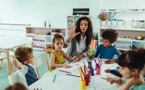 Une animatrice explique une activité créative à des enfants autour d'une table. Son rôle est d'éveiller leur curiosité.