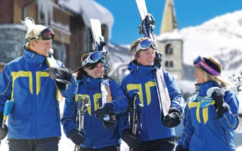 Quatre jeunes skieurs souriants en tenue bleue et jaune, portant des skis et des bâtons, dans une station de ski enneigée.