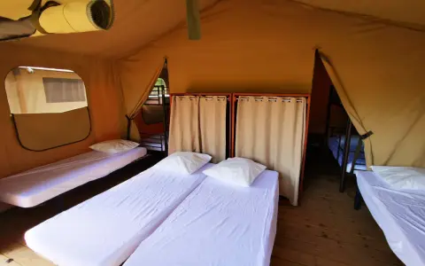 Intérieur d'une chambre de colonie de vacances avec plusieurs lits simples, draps blancs et oreillers. Des rideaux séparent les espaces.