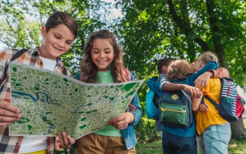 Deux enfants souriants regardent une carte dans un parc, d'autres enfants s'enlacent en arrière-plan.