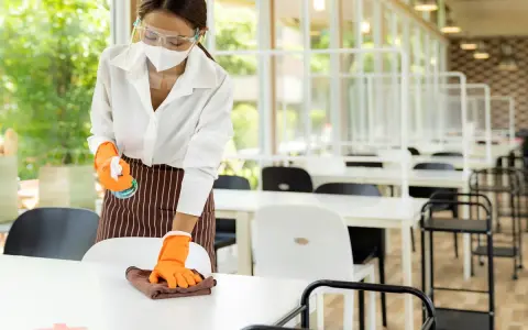 Employée portant masque et visière nettoie une table, respectant la réglementation hygiène cantine scolaire. Des séparateurs sont installés.