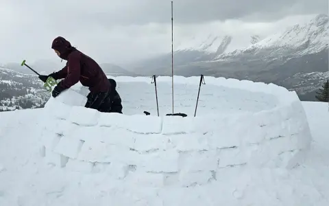 Person construisant un igloo en blocs de neige, avec des bâtons de ski plantés.