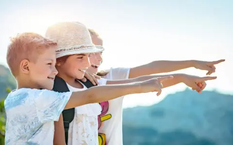 Trois enfants souriants, portant des sacs à dos, pointent du doigt vers la droite, sous un ciel ensoleillé.