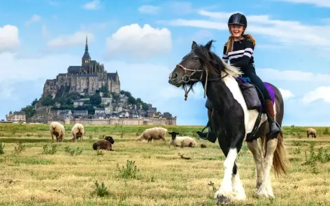 Une jeune fille rit à cheval devant le Mont-Saint-Michel, avec des moutons dans un pré.