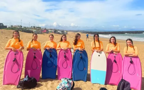 Huit jeunes filles en t-shirts orange, prêtes pour leur colonie à la mer, posent avec leurs bodyboards sur la plage.