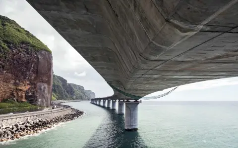 Vue sous un pont en béton, avec la mer, des falaises verdoyantes et une route côtière.