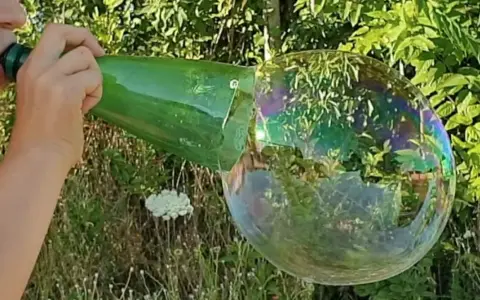 Main d'enfant soufflant dans une bouteille verte pour créer une bulle de savon géante irisée.