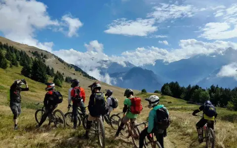 Groupe d'ados en colonie VTT descente, prêts à dévaler les pentes montagneuses sous un ciel bleu éclatant.