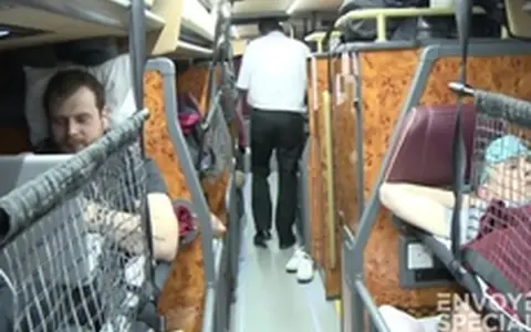 Intérieur d'un bus avec des couchettes. Un homme en blanc marche dans l'allée, tandis que des passagers se reposent dans leurs couchettes.