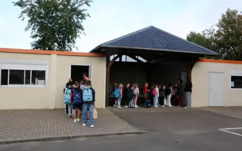 Des enfants font la queue devant un bâtiment scolaire, certains avec des sacs à dos.
