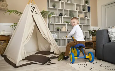 Un petit garçon sur un tricycle bleu et jaune s'amuse dans un espace de jeux, près d'une tente tipi et d'un canapé. Ce centre de vacances et de loisirs est idéal pour les enfants.