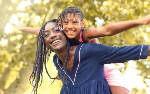 Une femme porte une enfant sur son dos, toutes deux souriantes, avec des arbres en arrière-plan.