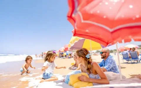 Famille souriante sur une plage ensoleillée, sous des parasols colorés, avec des enfants jouant dans l'eau.