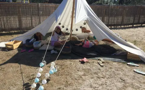 Enfants profitant d'une colonie de vacances camping dans une tente tipi, entourés de coussins et de guirlandes lumineuses.