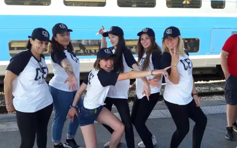 Groupe de jeunes femmes souriantes portant des casquettes et t-shirts "CEI 1947" devant un train.