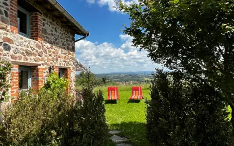 Maison en pierre avec vue panoramique sur les collines, deux chaises longues rouges invitent à la détente. Parfait pour découvrir les destinations Rhône-Alpes.