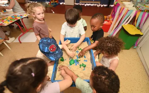 Des enfants s'amusent dans un bac à sable au centre de loisirs. Un petit garçon porte une casquette Spiderman. Ils ont tous environ 2 ans.