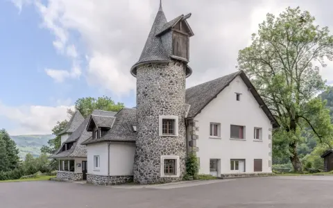 Bâtiment blanc avec une tour ronde en pierre et un toit en ardoise, entouré d'arbres et de verdure.