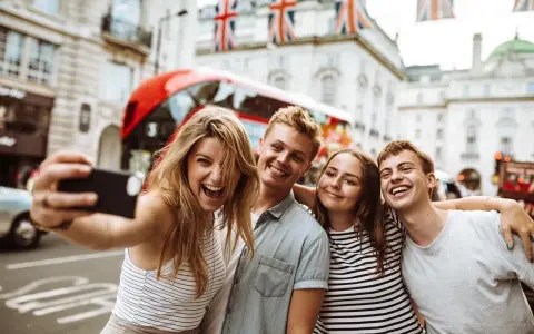 Quatre jeunes souriants prennent un selfie à Londres, avec un bus rouge et des drapeaux britanniques en arrière-plan.