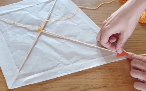 Mains fabriquant un cerf-volant en papier blanc avec des bâtons de bois et de la ficelle.