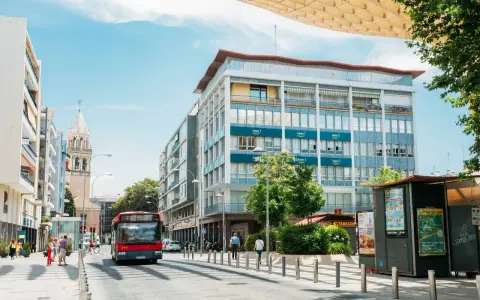 Un bus rouge circule dans une rue bordée d'immeubles et d'un clocher, sous un ciel bleu.