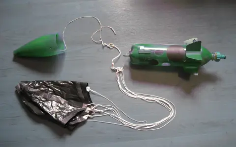 Fusée à eau verte avec cône, parachute noir et fils blancs sur fond gris.